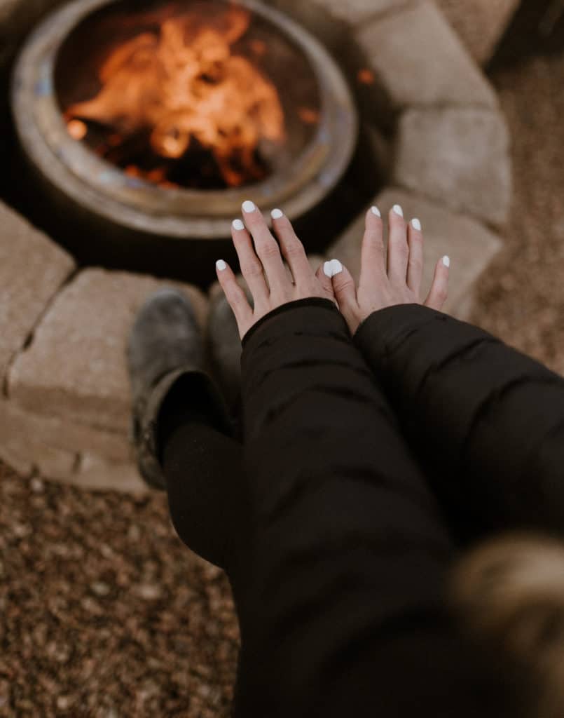 Girl Warms Hands Over Campfire Wearing a Black Jacket and White Nail Polish